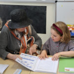 Two people (an elderly woman in a black hat and a young girl in a checkered shirt) examine worksheets at a table in a classroom, pointing at activity pages together.