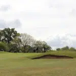 Group of golfers standing and walking on a golf course near a sand bunker with trees in the background under a cloudy sky.