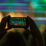 Person holds a smartphone to record a live concert, with a colorful, blurred stage behind them and silhouettes of the crowd in front.